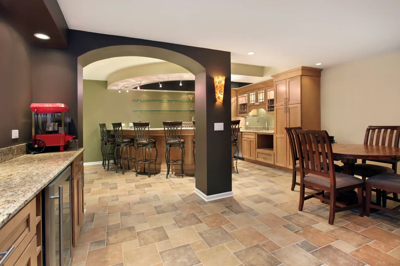Stylish home bar with wooden cabinets, high chairs, and a granite countertop. A red popcorn machine adds a fun touch. Warm, cozy ambiance.