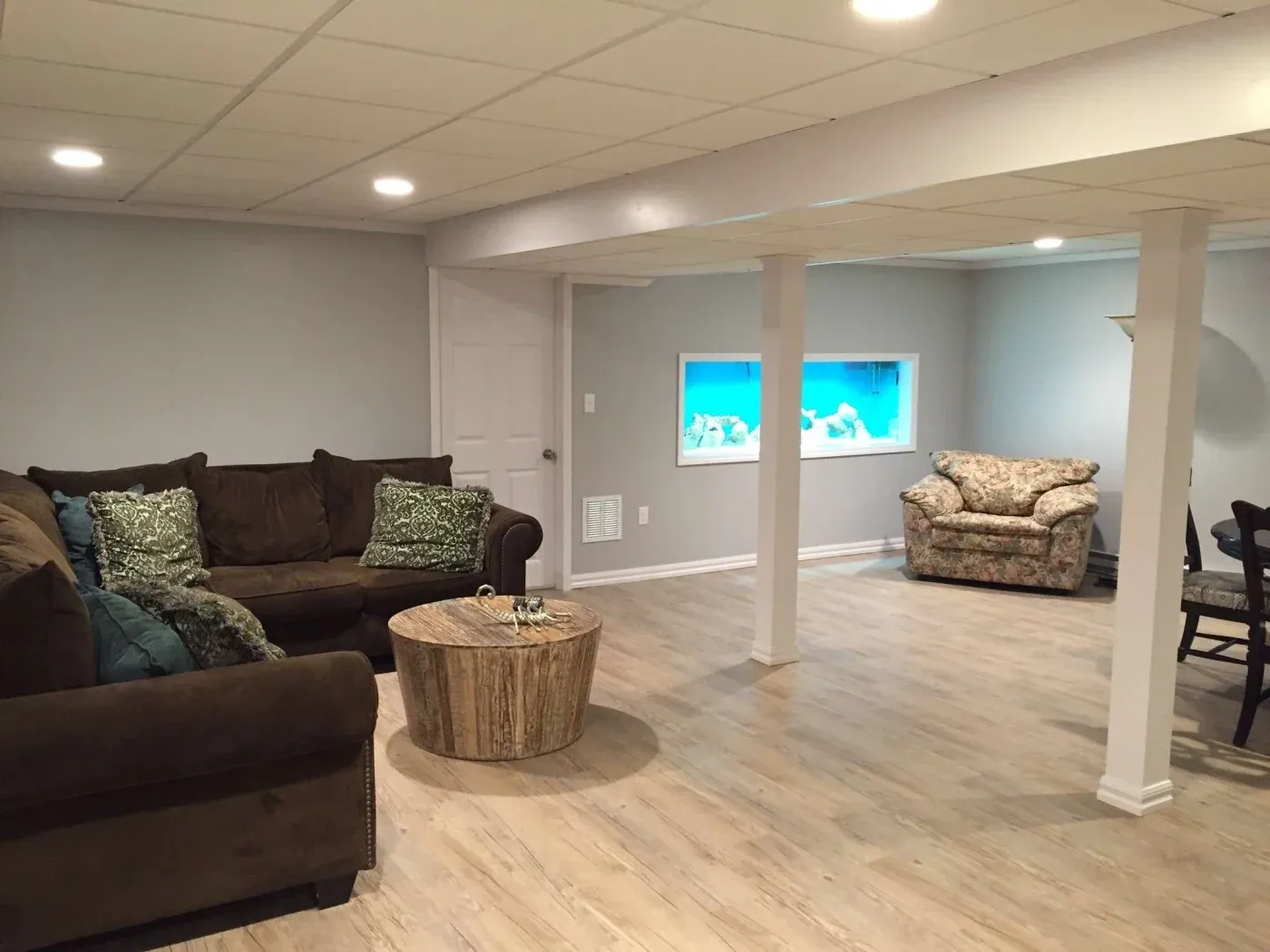 Spacious basement with light wood flooring, a brown sectional sofa, and floral armchair. Wall-mounted fish tank adds a serene, cozy atmosphere.