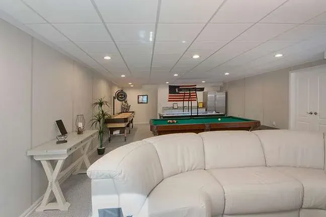 A modern games room with a white sectional sofa in the foreground, a pool table, a foosball table, and an American flag on the back wall.