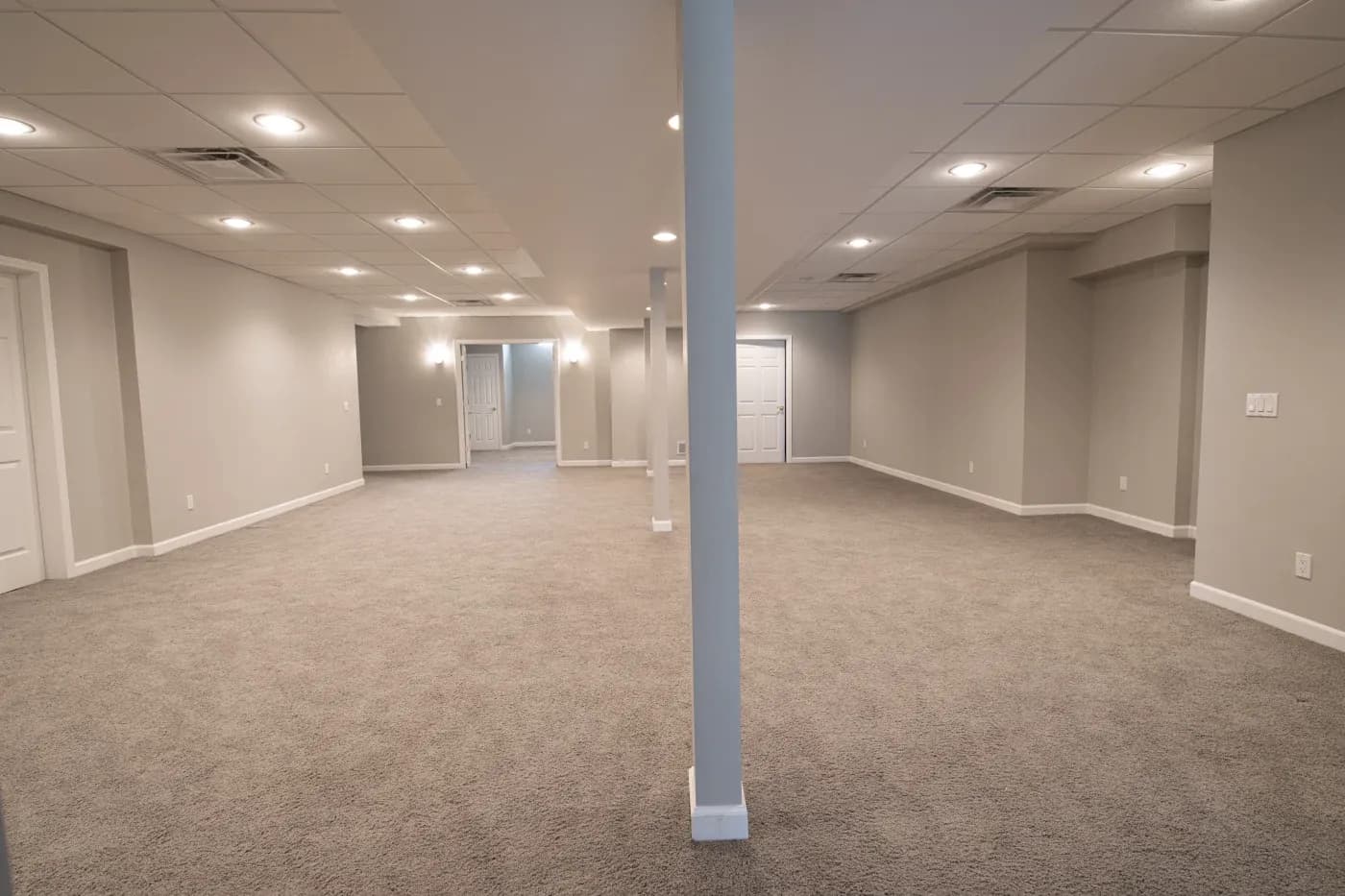 Spacious, empty basement with gray carpet, neutral walls, and bright ceiling lights. Two support columns divide the area. Doors are visible in the back.