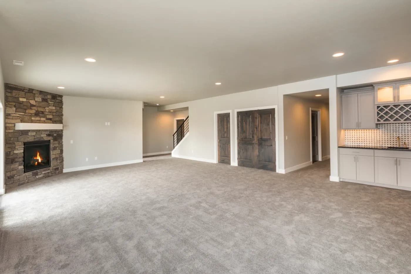 Spacious modern living room with stone fireplace, plush gray carpet, recessed lighting, wooden doors, and a built-in kitchenette with wine rack.