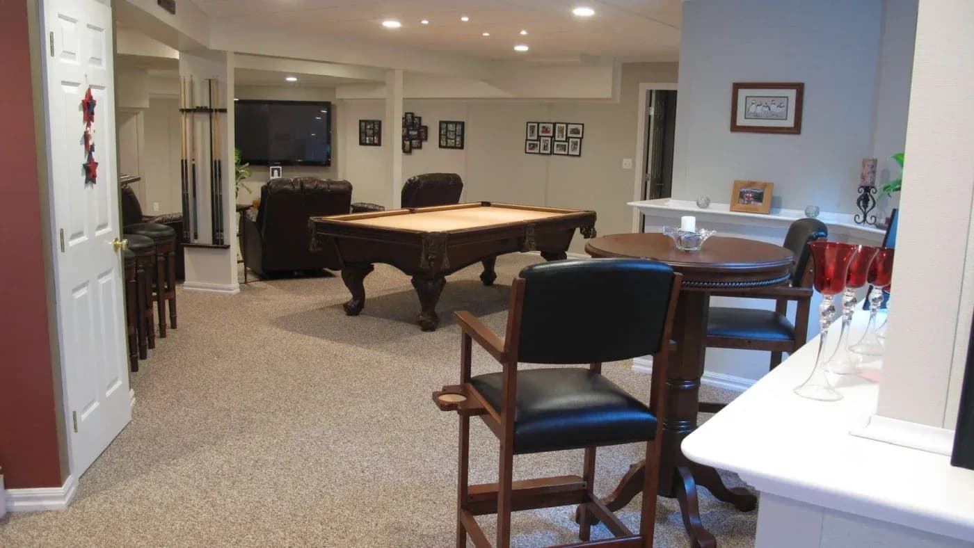 A cozy basement game room features a pool table, leather sofas, and a wall-mounted TV. Bar stools and framed photos create a welcoming atmosphere.