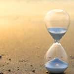 A small hourglass with white sand passing through it sits on a nondescript dark surface with sunlight flaring from the left-hand background