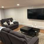 A finished basement media room with dark gray recliners, wood-look flooring, recessed lighting, a large wall-mounted TV, and a patterned area rug.