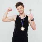 A man flexing while he has a gold medal around his neck and holding a gold trophy.