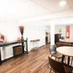 Open-concept basement finishing project with wood-look flooring, a round dining table, leather seating, recessed lighting, and an entertainment area with a mounted TV and wet bar.