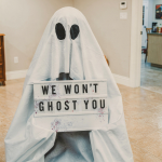 A person in a ghost outfit holding a sign that says “We Won’t Ghost You” while standing in a newly finished basement.