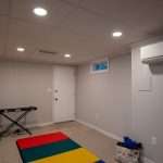 The lower level of a home has been turned into a multi-purpose space, with a colorful mat on the floor, a keyboard and guitar in front of the far wall, and a tote bag next to the mat. The walls are light gray in color, and the carpet is tan.