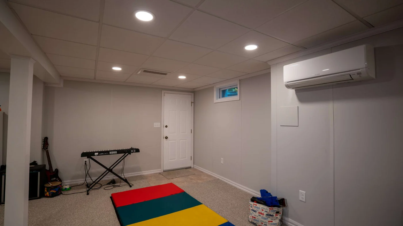 The lower level of a home has been turned into a multi-purpose space, with a colorful mat on the floor, a keyboard and guitar in front of the far wall, and a tote bag next to the mat. The walls are light gray in color, and the carpet is tan.