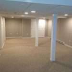 A photo shows the finished lower level of a home, complete with light gray paneled walls, tan carpet, and white doors, trim, and support columns.