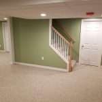 A portion of a lower level finished with pale tan panels on the side walls, and olive green on an accent wall. The carpet is light colored, and the trim and doors are white, along with the ceiling. The lower part of a staircase with a brown wooden railing is visible along the far wall.